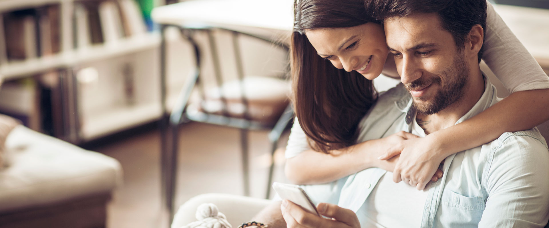 Cox & Co Estate agent in Edinburgh, Scotland. A woman with long brown hair embraces a man with a beard from behind as they both smile and look at a smartphone he's holding. They are sitting in a cozy, warmly-lit living room, possibly discussing details with their mortgage broker, with shelves and a chair in the background.