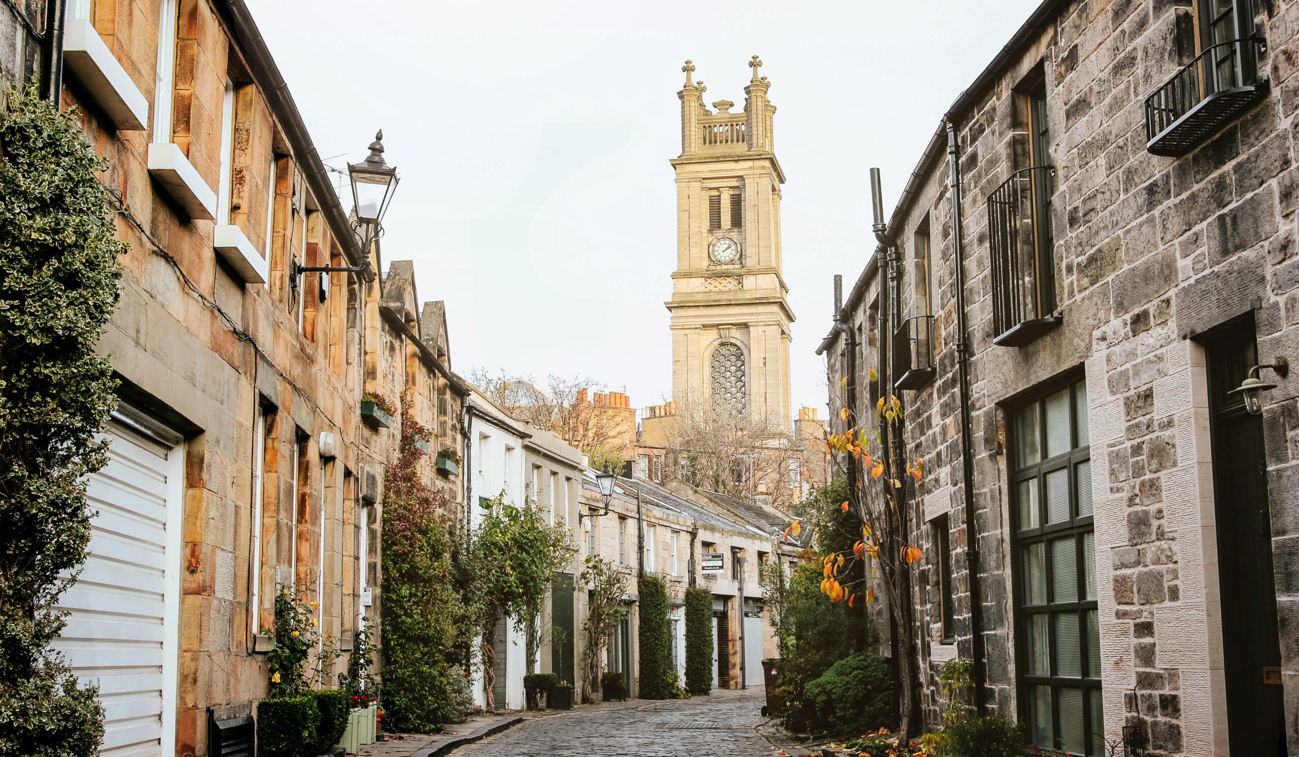 Cox & Co Estate agent in Edinburgh, Scotland. A narrow, cobblestone street in one of Edinburgh's best areas is lined with stone buildings and leafy greenery, leading to a tall church tower with a clock near the top. The tower is centered in the background, while the street and buildings create a quaint, historic ambiance perfect for those looking to buy property.