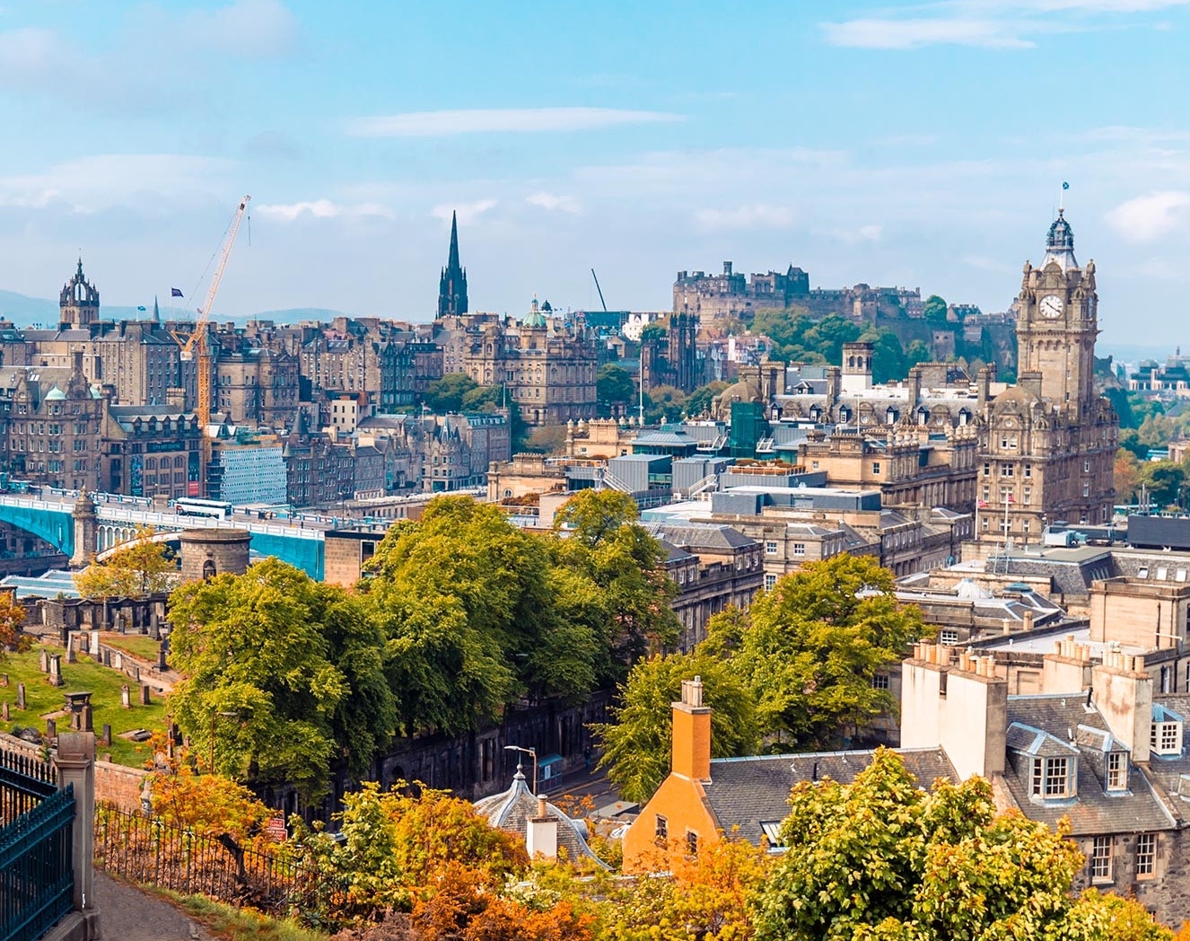 Cox & Co Estate agent in Edinburgh, Scotland. A panoramic view of Edinburgh, Scotland, showcasing historic buildings, green trees, and the prominent Edinburgh Castle in the background. The cityscape features a mix of old and newer architecture under a partly cloudy blue sky—a true gem for property investment.