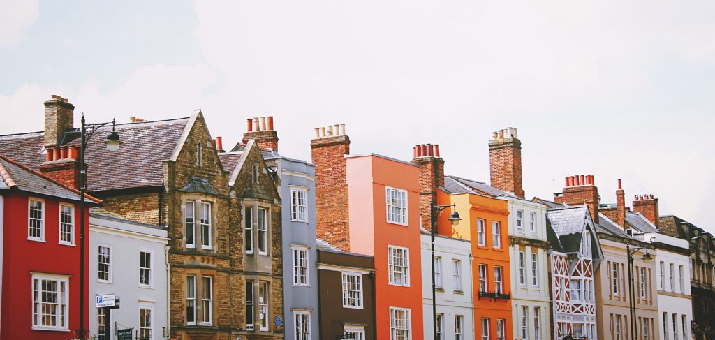 Cox & Co Estate agent in Edinburgh, Scotland. A row of colorful terraced houses in a quaint neighborhood showcases a variety of architectural styles, perfect for those interested in property investment. The buildings feature vibrant colors like red, orange, and gray with brick chimneys and large windows. The sky above is bright and slightly cloudy.