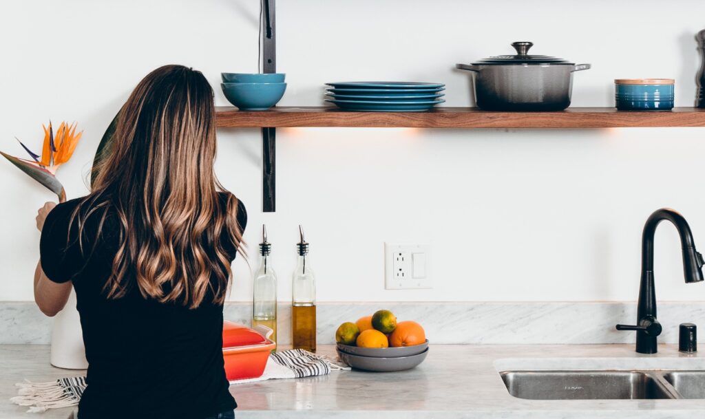 Cox & Co Estate agent in Edinburgh, Scotland. A woman with long, wavy hair, wearing a black shirt, arranges a large flower in a white vase on a kitchen counter. The counter holds a bowl of citrus fruits, olive oil bottles, and a red dish. Above her is evidence of thoughtful property management—a wooden shelf with neatly stacked dishes and cookware.