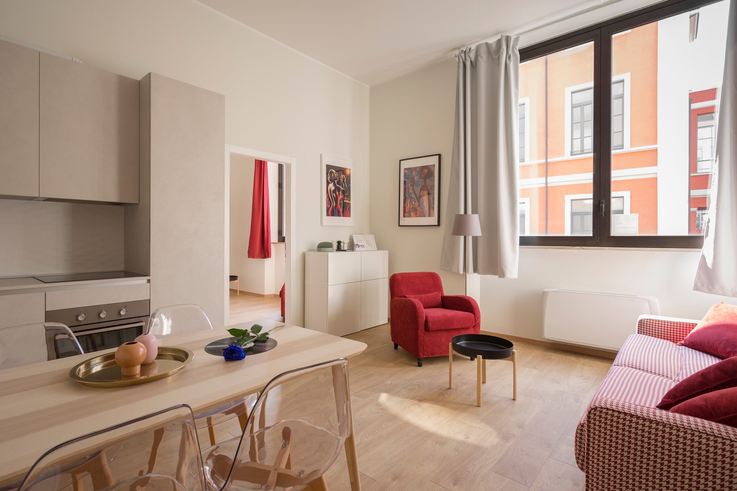 Cox & Co Estate agent in Edinburgh, Scotland. A bright and cozy living space featuring a red armchair, a striped red sofa, and a glass coffee table. The room has large windows with white curtains, framed art on the walls, a small kitchen area with light wooden cabinetry, and a dining table with transparent chairs—perfect for generating rental income.