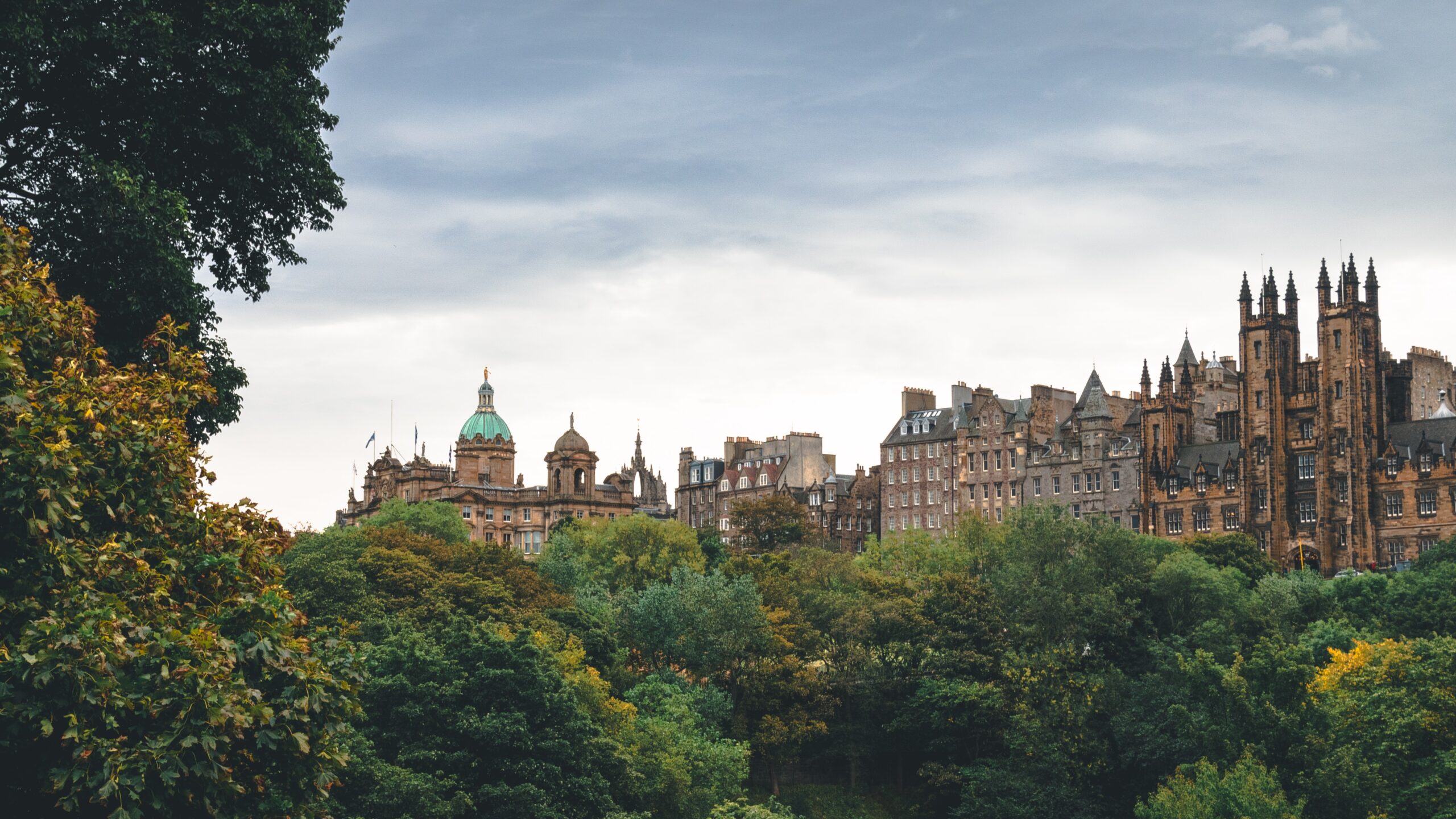 Cox & Co Estate agent in Edinburgh, Scotland. A scenic view of Edinburgh, Scotland, featuring historic buildings with spires and domes, partially obscured by lush green trees, under a cloudy sky. The architecture includes a mix of classic and gothic styles, making it an attractive prospect for those interested in investing in UK property.