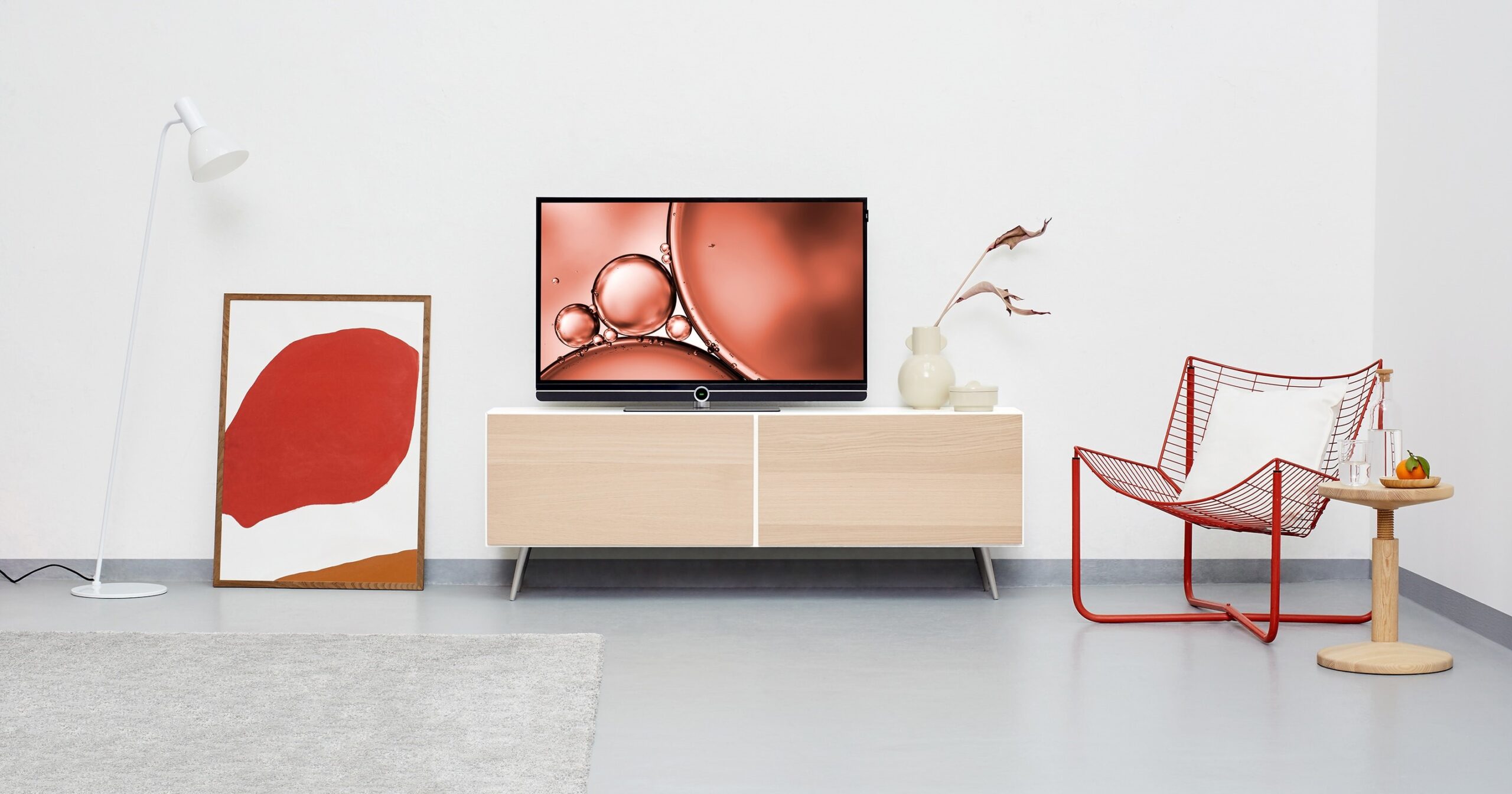 Cox & Co Estate agent in Edinburgh, Scotland. A modern living room featuring a light wooden TV stand with a flat-screen TV on top displaying an abstract image. A red wire chair with a white cushion and wooden side table are on the right, while a lamp and abstract red art piece are on the left—perfect for those looking to self-manage property effectively.
