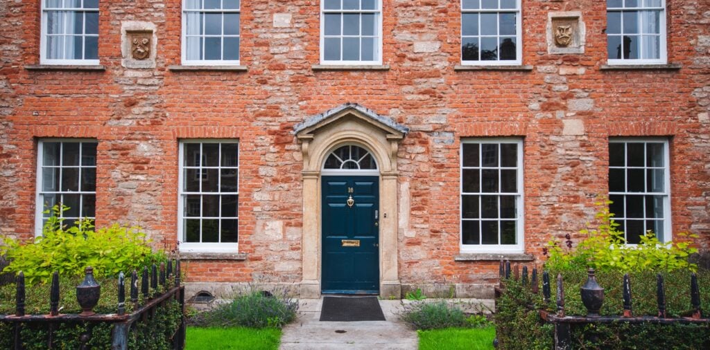 Cox & Co Estate agent in Edinburgh, Scotland. A red brick building with five large windows, a central arched doorway with a dark green door, and two stone plaques above the top windows. The front yard features a small garden with green shrubs and an iron fence, offering an ideal investment opportunity for high-net-worth individuals seeking million pound mortgages.