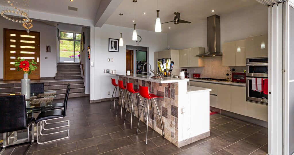 Cox & Co Estate agent in Edinburgh, Scotland. A modern kitchen with a stone-accented island, red high chairs, and stainless steel appliances. To the left is a dining area with a glass table and black chairs. In the background, steps lead to an open front door. Pendant lights hang from the ceiling, exemplifying ideal details for property investment.
