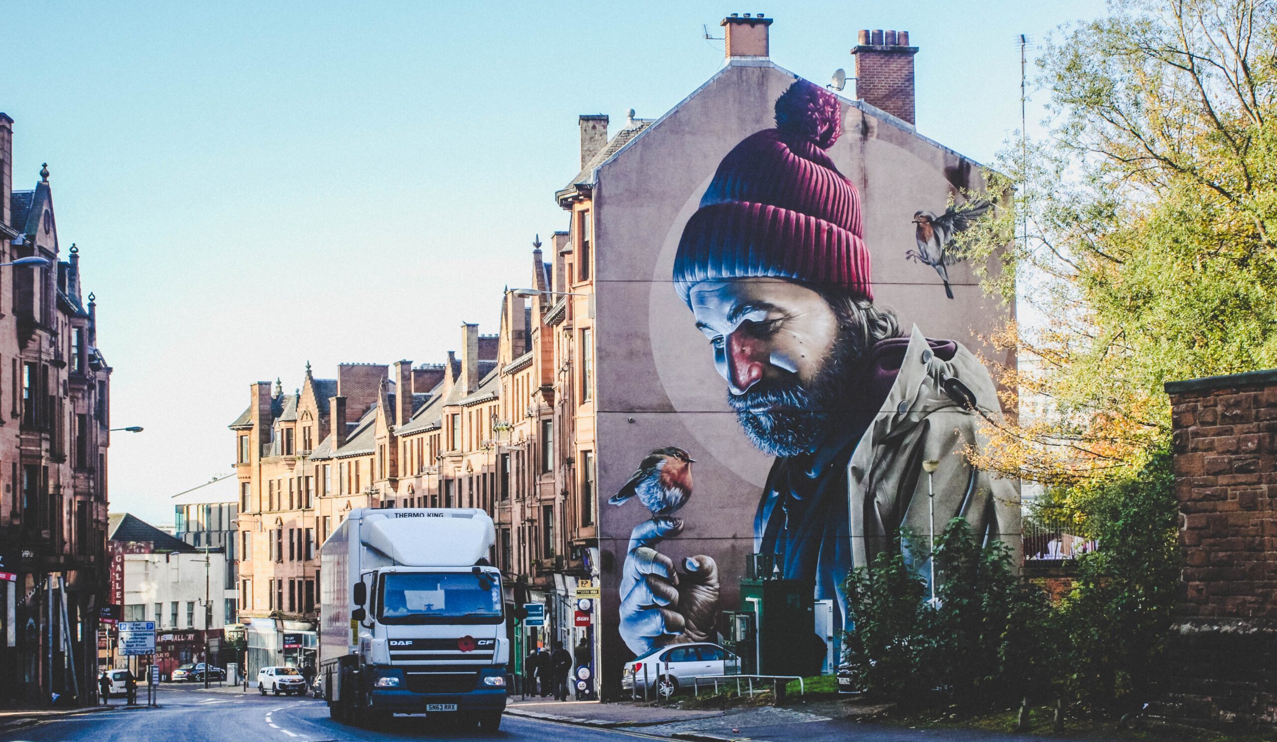 Cox & Co Estate agent in Edinburgh, Scotland. Street scene featuring a tall building with a large mural of a bearded man in a beanie holding a bird. A white truck drives past, and traditional buildings line the street under a clear blue sky. Trees on the right add a touch of greenery, making this area potentially right for you to consider investing.