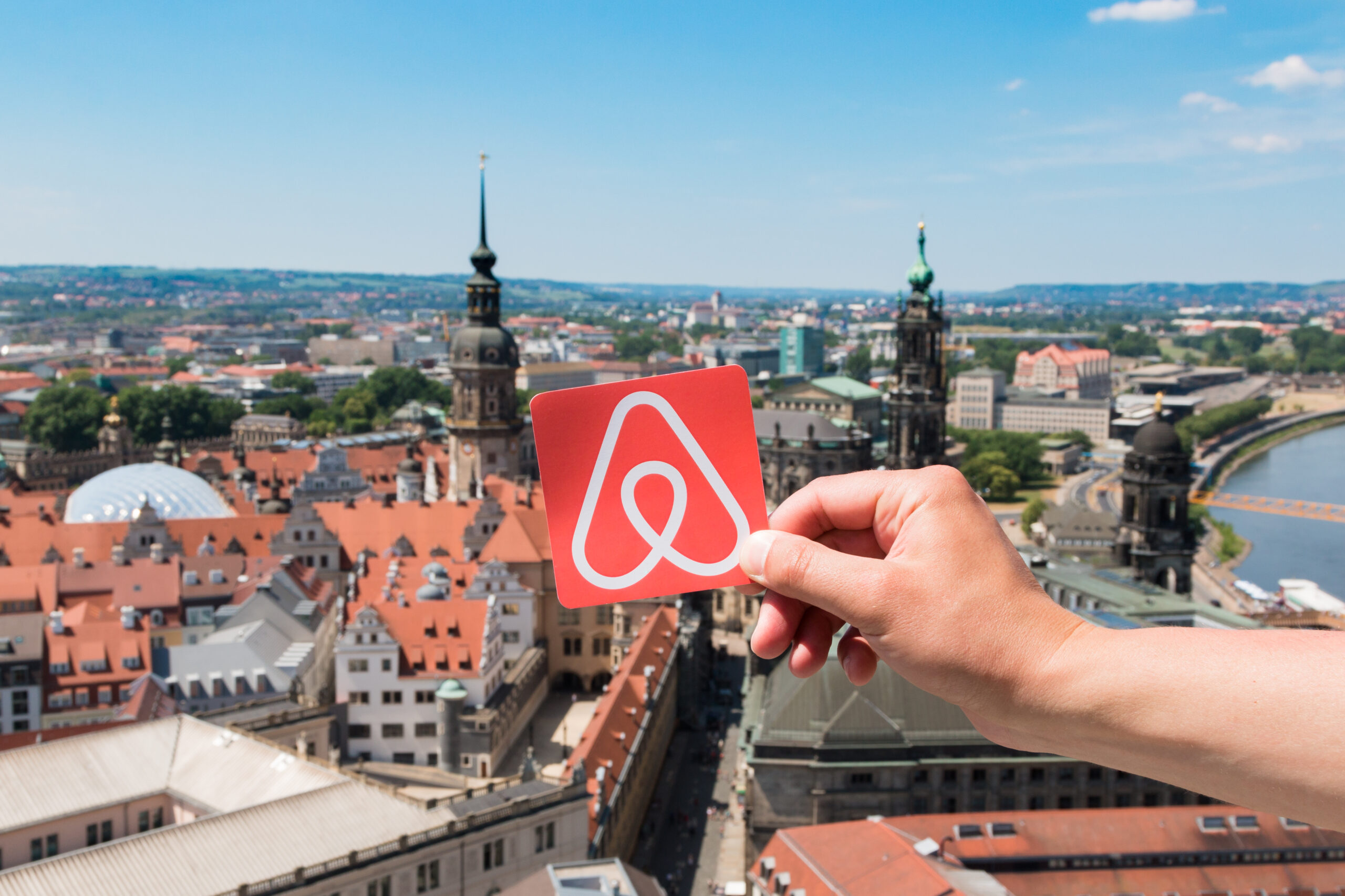 Cox & Co Estate agent in Edinburgh, Scotland. A hand is holding a red card with the Airbnb logo in front of a panoramic view of a historic European city with river, spires, and rooftops. The scene showcases architectural landmarks and a distant landscape under a clear sky—perfect reasons to think again about starting an Airbnb.