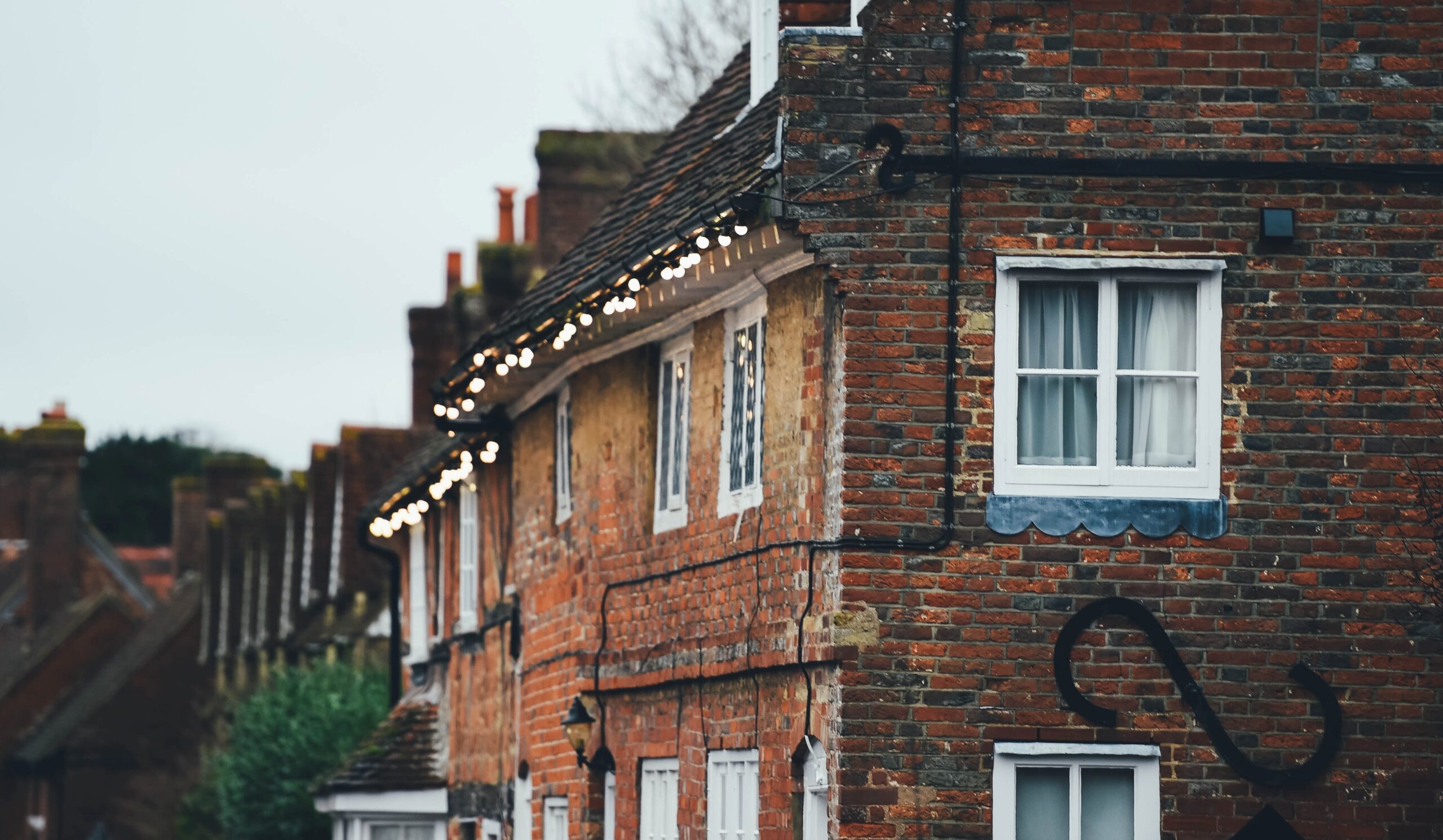 Cox & Co Estate agent in Edinburgh, Scotland. A row of old brick houses with white-trimmed windows and sloped roofs, adorned with hanging string lights, epitomizes UK property charm. The houses have visible chimneys, and greenery can be seen in the background under an overcast sky—a picturesque scene perfect for a buying guide for any US citizen.