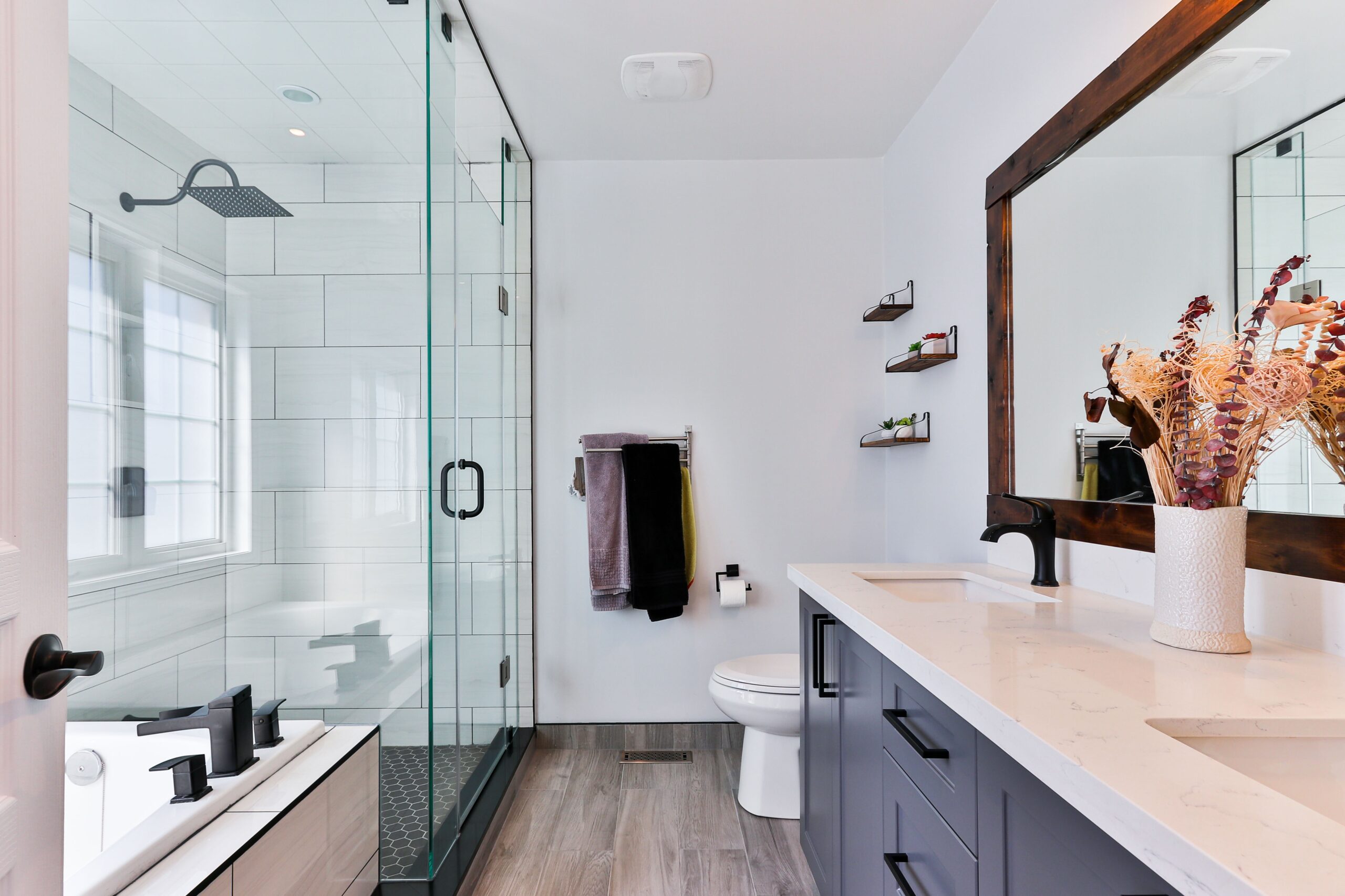 Cox & Co Estate agent in Edinburgh, Scotland. A modern bathroom that could increase property value, featuring a glass shower with a black overhead rain showerhead, a white bathtub, a toilet, and a vanity with a white countertop and black fixtures. Dried flowers in a vase decorate the vanity beside a large framed mirror, with floating shelves on the wall.