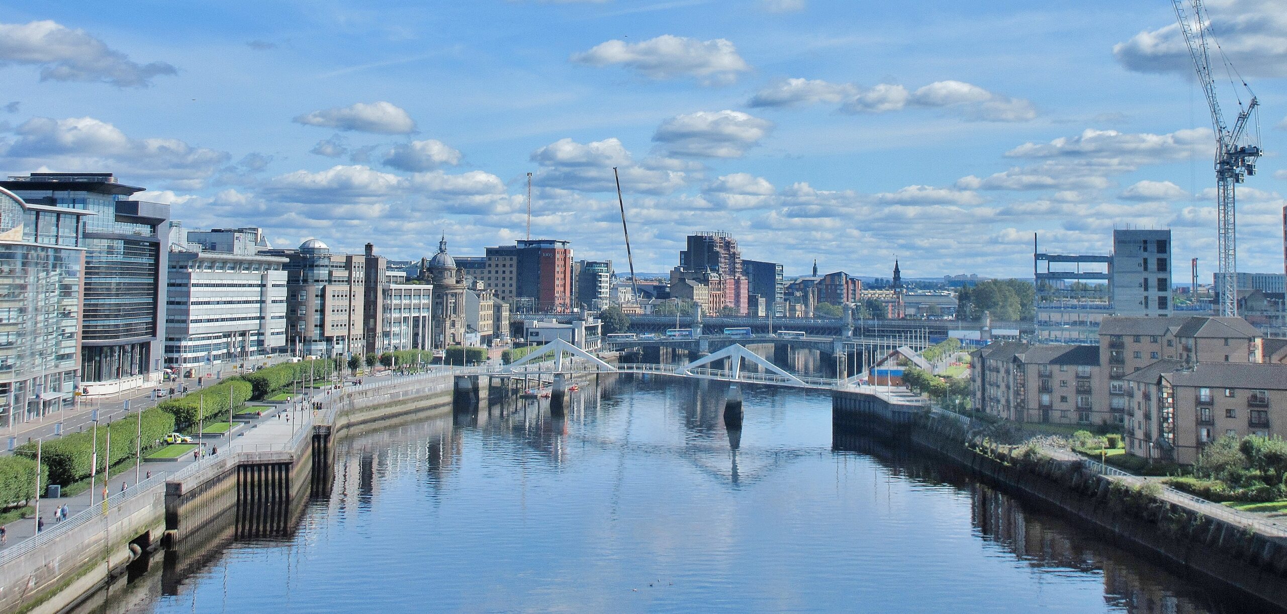 Cox & Co Estate agent in Edinburgh, Scotland. A vibrant cityscape featuring a river running through the center, bordered by modern and historic buildings. Pedestrian walkways and greenery line the riverbanks. Several bridges span the water, with a crane and construction visible in the background under a partly cloudy sky—showcasing 2021 Glasgow Property Investment opportunities.
