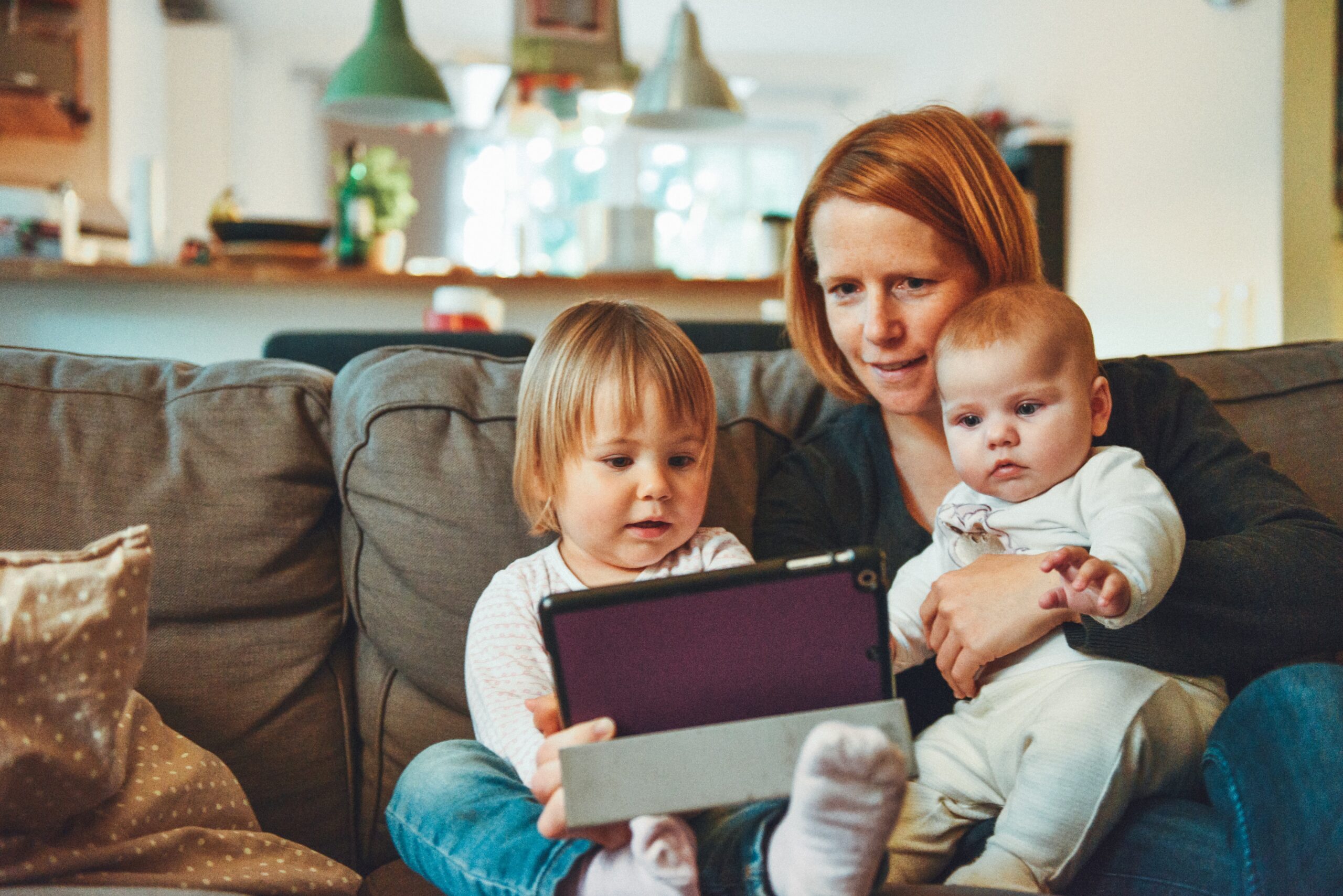 Cox & Co Estate agent in Edinburgh, Scotland. A woman with red hair sits on a couch with two young children. The older child, wearing light-colored pajamas, holds a tablet. The baby, also dressed in light-colored clothes, is on the woman's lap. They all focus intently on the screen, much like perfect tenants meticulously selected for an investment property.