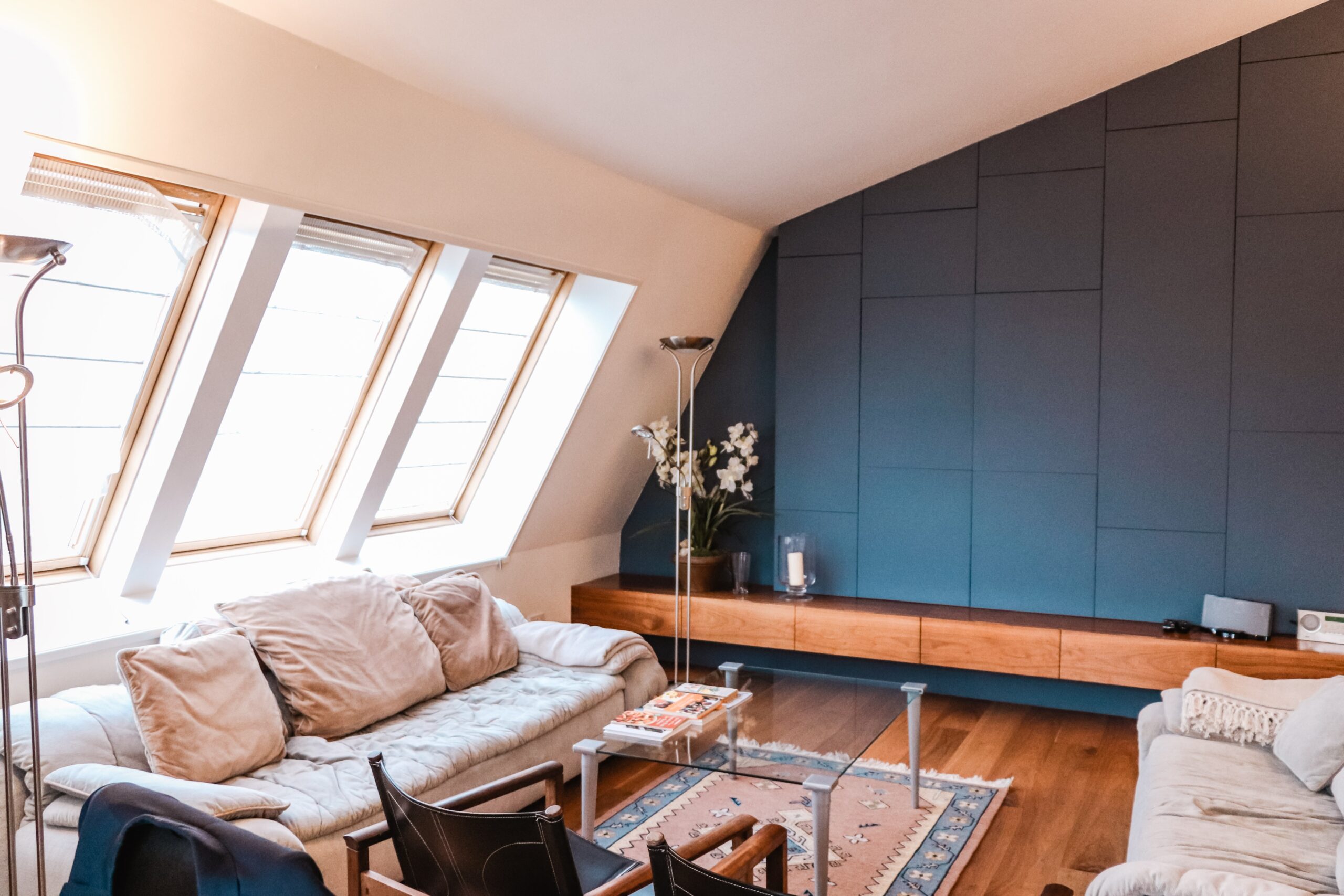 Cox & Co Estate agent in Edinburgh, Scotland. A modern living room with large slanted windows on the left, white walls, a dark blue accent wall, and wooden floors. Ideal for an investment property or new property, the space features beige sofas, a glass coffee table, a wooden floating console, and a floor lamp. A plant and books decorate the area.