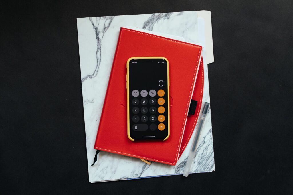 Cox & Co Estate agent in Edinburgh, Scotland. A smartphone displaying a calculator app rests on top of a closed red notebook, ready to help you calculate ROI for your investment property. Beneath the notebook is a folder with a marble-like pattern, and to its right lies a pen, all set against a dark background.