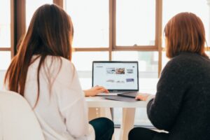 Cox & Co Estate agent in Edinburgh, Scotland. Two women seated at a table in a brightly lit room, viewing a laptop screen together. The laptop displays a website with various links and images. They appear to be collaborating or discussing tips for property managers related to the content on the screen.