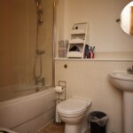 Cox & Co Estate agent in Edinburgh, Scotland. A small bathroom with a bathtub-shower combo on the left, enclosed by a transparent glass door. A toilet and a pedestal sink are on the right, with a circular mirror above the sink. Various toiletries are stored on white shelves above the toilet and sink, creating an organized court for your essentials.