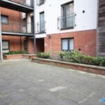 Cox & Co Estate agent in Edinburgh, Scotland. A modern apartment complex with red brick and white exterior walls. A large, paved courtyard features a raised garden bed with various plants and shrubs. Metal balconies and railings extend from the building. Small windows are visible on the lower level, adding charm to the court-like setting.