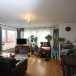 Cox & Co Estate agent in Edinburgh, Scotland. A spacious, bright living room with wooden floors, a brown leather couch, and a TV on a stand by the window. Various potted plants, toys, and a bookshelf contribute to the cozy feel. A clock hangs on the wall like a silent court judge, while sheer curtains filter in light.