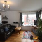 Cox & Co Estate agent in Edinburgh, Scotland. A living room with a leather sofa, coffee table, TV on a wooden stand, and large windows covered with sheer curtains. A decorative plant stands in the corner like a watchful courtier. A colorful toy mat lies on the wooden floor, scattered with various children’s toys like a playful salamander's treasure trove.
