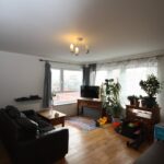 Cox & Co Estate agent in Edinburgh, Scotland. A well-lit living room in Salamander Court with wooden floors, a brown leather sofa, a coffee table, and children's toys on a colorful rug. The room features large windows with curtains, a TV on a stand, assorted plants, a bookshelf, and a ceiling light fixture.