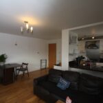 Cox & Co Estate agent in Edinburgh, Scotland. A living room at Salamander Court with wooden flooring features a black leather couch and a small office area with a folding chair and desk. Ceiling lights illuminate the room, revealing a partially visible kitchen area with modern appliances. A few houseplants add greenery to the space.