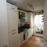 Cox & Co Estate agent in Edinburgh, Scotland. A modern kitchen at Salamander Court features white cabinets, wooden countertops, and stainless steel appliances. There's a gas stove with a range hood, fridge, and small oven. Natural light streams in through a window with sheer floral curtains, leading to a cozy balcony with plants.