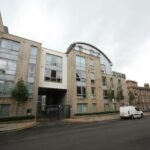 Cox & Co Estate agent in Edinburgh, Scotland. A modern apartment building on Gardners Crescent features large windows and a mix of beige stone and gray paneling. The structure includes multiple levels and a curved roof section. A white van is parked on the street in front, and a gated entrance is visible on the ground floor.