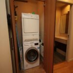 Cox & Co Estate agent in Edinburgh, Scotland. A small laundry nook with a stacked washer and dryer is visible between partially open wooden doors. A cleaning cloth and some supplies are on top of the dryer. To the side, there is a mop and an ironing board. In the background, a bathroom at Gardners Crescent is visible.