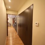 Cox & Co Estate agent in Edinburgh, Scotland. A hallway with light wood flooring, beige walls, and recessed ceiling lights. On the right, there's a dark wood storage cabinet with multiple doors and a thermostat on the wall. The hallway leads to an open kitchen area with modern appliances visible, reminiscent of the style found on Gardners Crescent.