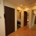 Cox & Co Estate agent in Edinburgh, Scotland. A hallway with light wooden flooring at Gardners Crescent has three open doors leading to different rooms. The walls are painted cream, and there is a dark wooden built-in wardrobe on the left. The area is well-lit with recessed ceiling lights.