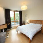 Cox & Co Estate agent in Edinburgh, Scotland. A bedroom on Gardners Crescent features a wooden double bed, a light comforter, and two bedside tables with lamps. The room has a desk and chair near a large window with dark curtains. A small laundry basket is placed next to the desk. The wooden floor and light-colored walls complete the cozy setting.
