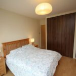 Cox & Co Estate agent in Edinburgh, Scotland. A cozy bedroom on Gardners Crescent with light-colored walls and wooden flooring. The room features a wooden bed with a floral bedspread, flanked by two wooden nightstands each with a lamp. A large wardrobe with sliding wooden doors is against one wall. A ceiling light hangs above.
