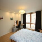 Cox & Co Estate agent in Edinburgh, Scotland. A minimalist bedroom on Gardners Crescent features a large bed with a patterned duvet cover, a desk with a black chair, and a table lamp. The room has hardwood floors, a large window with curtains, two small framed pictures on the walls, and a paper lantern hanging from the ceiling.