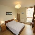 Cox & Co Estate agent in Edinburgh, Scotland. A tidy, well-lit bedroom on Gardners Crescent features a double bed centered against a wall. The bed has wooden side tables with lamps on either side. Above the bed is a framed picture. The room boasts a large window with a radiator beneath it, a wooden dresser, and a chair in the corner.
