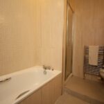Cox & Co Estate agent in Edinburgh, Scotland. A modern bathroom on Gardners Crescent featuring a white bathtub with chrome handles on the left, beige tiled walls, a glass-enclosed shower next to the bathtub, a heated towel rack with a beige towel, and a white toilet on the right. The space has a clean and minimalist design.