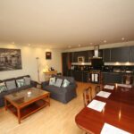 Cox & Co Estate agent in Edinburgh, Scotland. A well-lit, open-plan living area with a modern kitchen on Crescent. The space includes a dark wooden dining table set, a cozy seating area with a grey sofa and armchairs, a wooden coffee table, and wall art. The kitchen features dark cabinets, a built-in oven, and various appliances for all Gardners' needs.