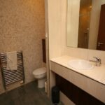 Cox & Co Estate agent in Edinburgh, Scotland. A modern bathroom on Gardners Crescent, with beige tiled walls and a dark tiled floor. It features a white sink with a stainless steel faucet on a beige countertop, a large mirror above, a toilet, a wall-mounted towel rail with a beige towel, and a black trash can underneath the sink.