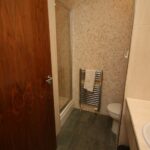 Cox & Co Estate agent in Edinburgh, Scotland. A small bathroom with a modern design features a shower with a glass door, a towel hanging on a heated towel rail, a toilet, and a sink with a beige countertop. The walls are tiled, and the floor has dark green tiles reminiscent of Gardners Crescent. The wooden door is partially open.