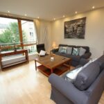 Cox & Co Estate agent in Edinburgh, Scotland. A well-lit living room with large windows on Gardners Crescent features a light wooden floor and modern furniture. The room includes two dark grey sofas, a wooden coffee table, a reading chair by the window, and a black-and-white cityscape picture on the wall.
