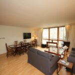 Cox & Co Estate agent in Edinburgh, Scotland. A spacious living room on Gardners Crescent with light wood flooring features a gray couch, a wooden coffee table, and a flat-screen TV mounted on the wall. There is a dining area with a wooden table and chairs. Large windows provide ample natural light, and various lamps brighten the space.