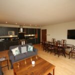 Cox & Co Estate agent in Edinburgh, Scotland. A spacious open-plan living area on Gardners Crescent with a kitchen, dining space, and lounge. The kitchen boasts dark cabinets and modern appliances; the dining area features a wooden table with chairs, while the lounge includes a sofa, armchairs, and a large wall-mounted TV. Light wood flooring ties it all together.