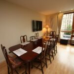 Cox & Co Estate agent in Edinburgh, Scotland. A dining room with a long wooden table set for eight people, each place adorned with a placemat. Chairs with carved backs surround the table. A wall-mounted flat-screen TV is visible, and a large window offers a view of the lush Gardners Crescent outside.