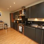 Cox & Co Estate agent in Edinburgh, Scotland. Modern kitchen on Gardners Crescent with dark cabinets, stainless steel appliances, and a black granite countertop. There is a stove with an overhead hood, a microwave, a sink, and a dish rack. The room has wooden flooring and warm lighting. A small wooden shelf and a trash bin are on the left.