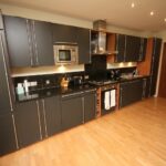 Cox & Co Estate agent in Edinburgh, Scotland. A modern kitchen on Gardners Crescent boasts sleek grey cabinets and a long black countertop. Stainless steel appliances include a microwave and stovetop with an overhead exhaust. Wooden flooring contrasts with the dark cabinetry, while bright lighting illuminates the space.