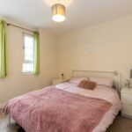 Cox & Co Estate agent in Edinburgh, Scotland. A cozy bedroom with a double bed covered in pink and white bedding. The bed is flanked by white nightstands, one with a gray lamp. Green curtains frame the two windows on the left wall. A ceiling light fixture hangs above the bed, perfect for those considering property investment.