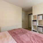 Cox & Co Estate agent in Edinburgh, Scotland. A bedroom with beige walls features a neatly made bed with pink and mauve bedding on the left. A white shelving unit with baskets and decorations stands at the foot of the bed. A vanity table with a mirror is on the right, and two closed white doors are in the back, perfectly staged by an estate agent in Edinburgh.