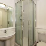 Cox & Co Estate agent in Edinburgh, Scotland. A small, modern bathroom with a glass-enclosed corner shower, a white pedestal sink, and a white toilet. The walls are light-colored, and a mirror hangs above the sink. The space is well-lit with ceiling lights—an ideal setting for those seeking premium property management solutions.