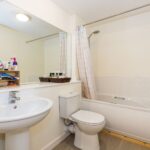 Cox & Co Estate agent in Edinburgh, Scotland. A clean and well-lit bathroom features a white sink with a chrome faucet, a mirror above it, a toilet, and a bathtub with a shower curtain on the right. Various toiletries are organized on a wooden shelf next to the sink. The design is simple and functional, perfect for an estate agent in Edinburgh to show potential buyers.