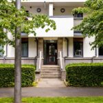 Cox & Co Estate agent in Edinburgh, Scotland. A residential building with a central entrance featuring a glass door and concrete steps. The facade is white with balconies, surrounded by lush green trees and well-maintained hedges—ideal for property investment. A sidewalk and a lamppost are visible in the foreground.