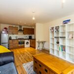 Cox & Co Estate agent in Edinburgh, Scotland. A modern, open-plan living room and kitchen with wooden flooring. The kitchen features brown cabinets, stainless steel appliances, including an oven, stove, and refrigerator. The living area includes a grey sofa, a yellow chair, a wooden coffee table, and a white shelving unit—ideal for property management.